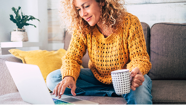 Woman in a yellow sweater sitting on a couch. She’s holding a coffee mug in one hand while scrolling on her laptop with the other.