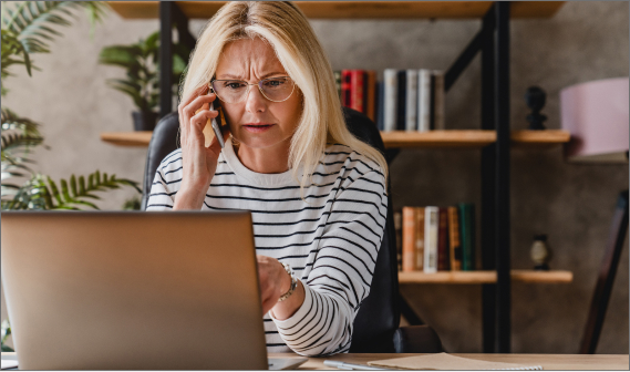 A woman is sitting at a desk. She is on her mobile phone and pointing to her laptop screen.